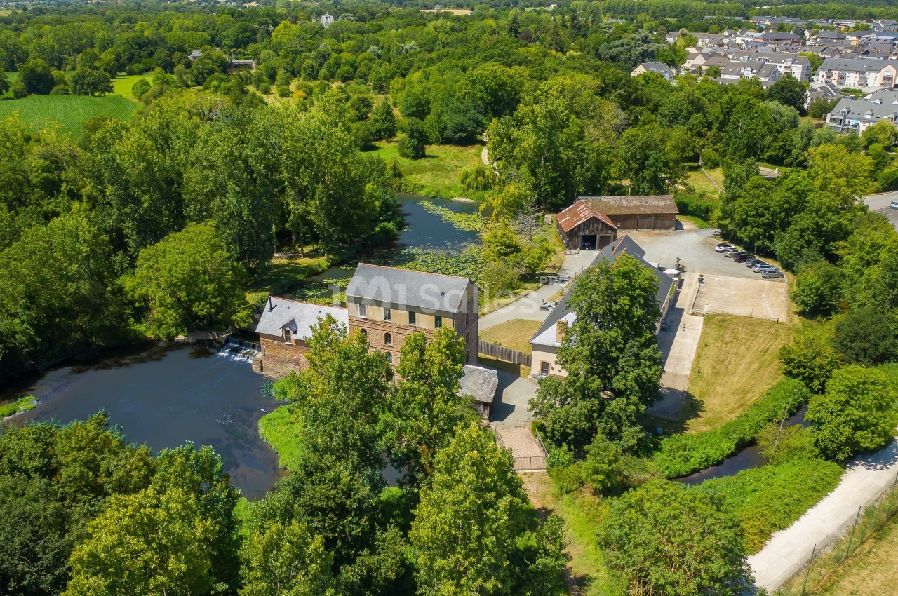 Vue aérienne d'un moulin en pierre entouré de verdure, avec une rivière et des bâtiments annexes à proximité.
