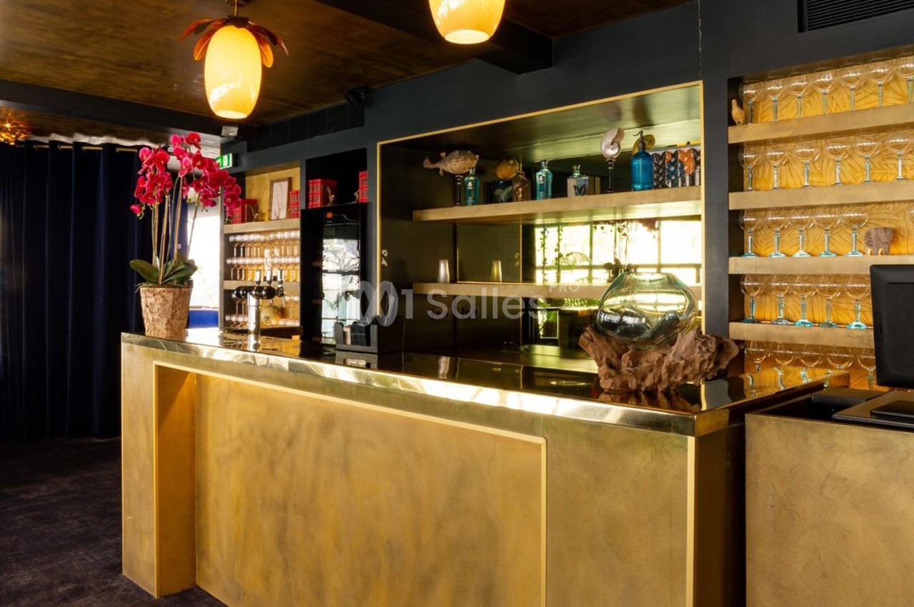 Comptoir doré dans un bar élégant avec étagères de verres, orchidée rouge et éclairage chaleureux.