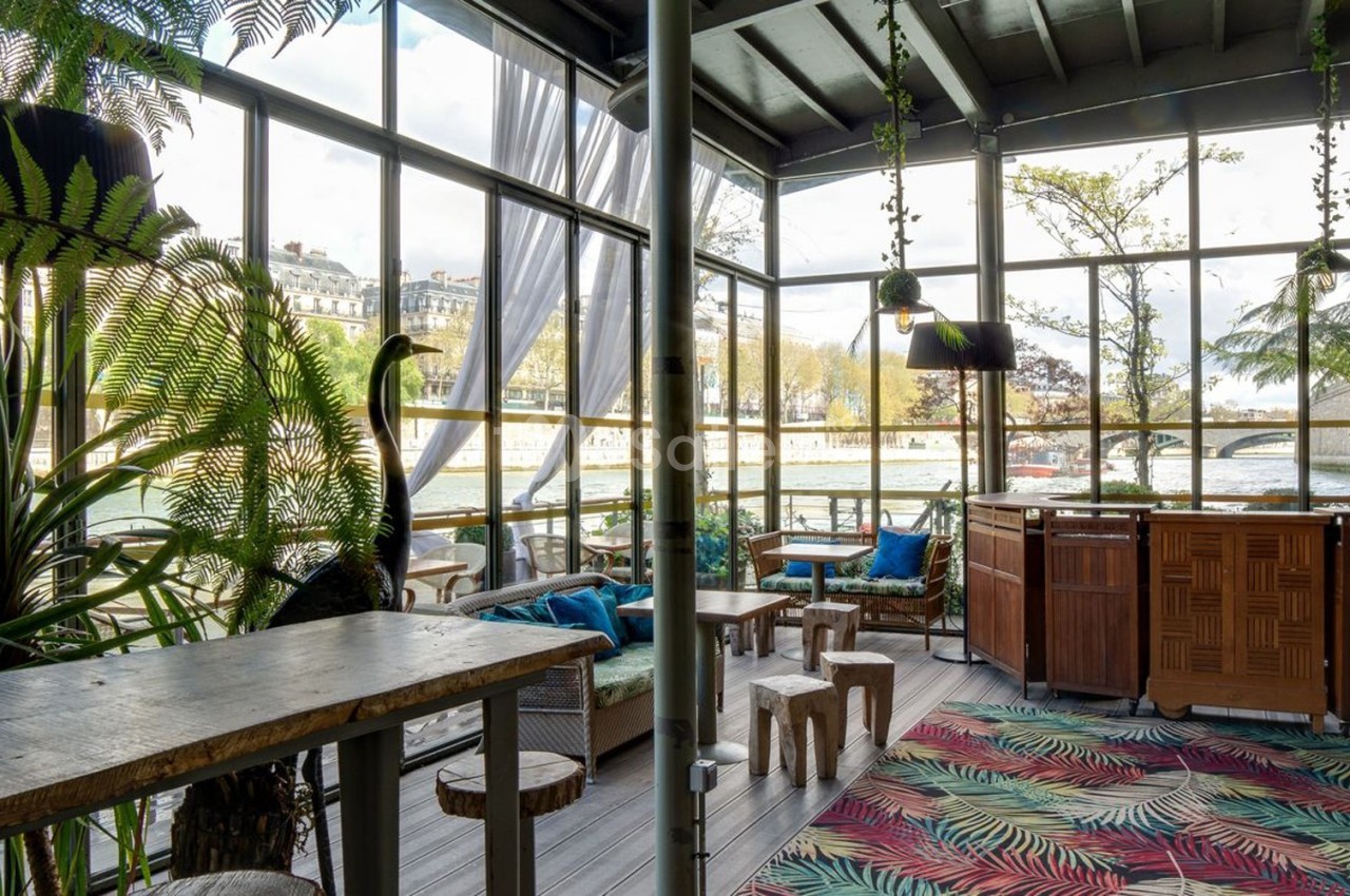 Intérieur lumineux d'une véranda avec mobilier en bois, plantes tropicales et vue sur un paysage urbain.