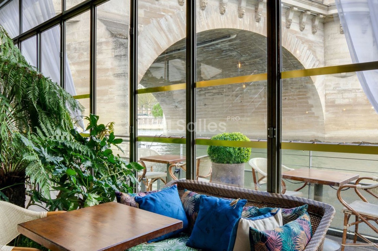 Espace de détente avec fauteuils et plantes, vue sur un pont en pierre à travers de grandes baies vitrées.