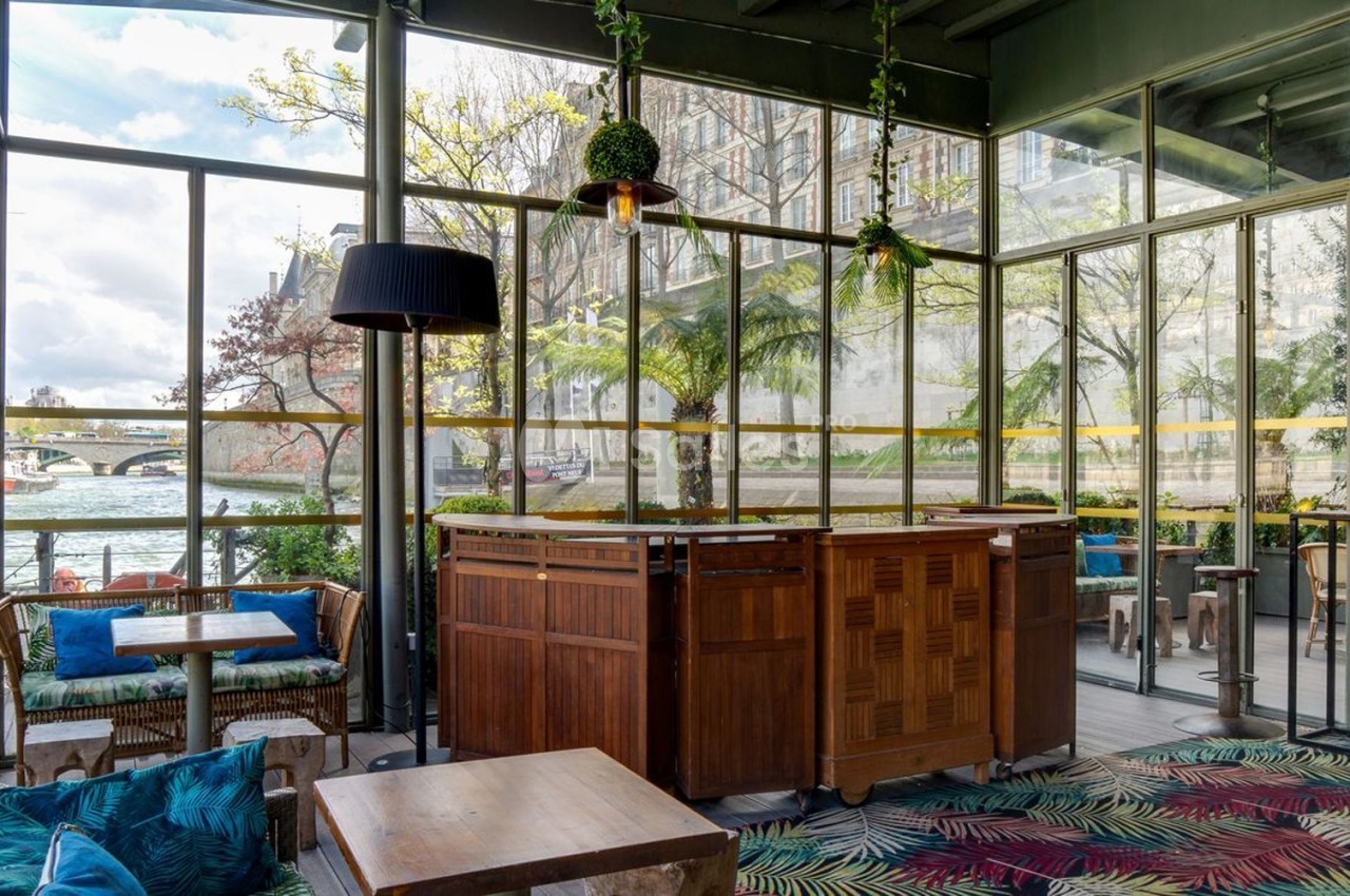Intérieur d'une véranda lumineuse avec mobilier en bois, plantes suspendues et vue sur un fleuve à travers de grandes baies…