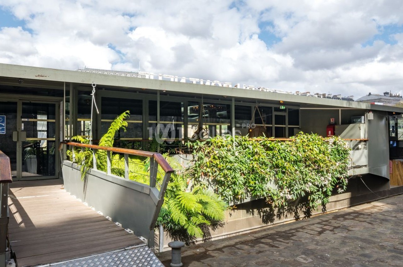 Bâtiment flottant moderne avec façade végétalisée, accessible par une passerelle en bois, amarré le long d'un quai pavé.
