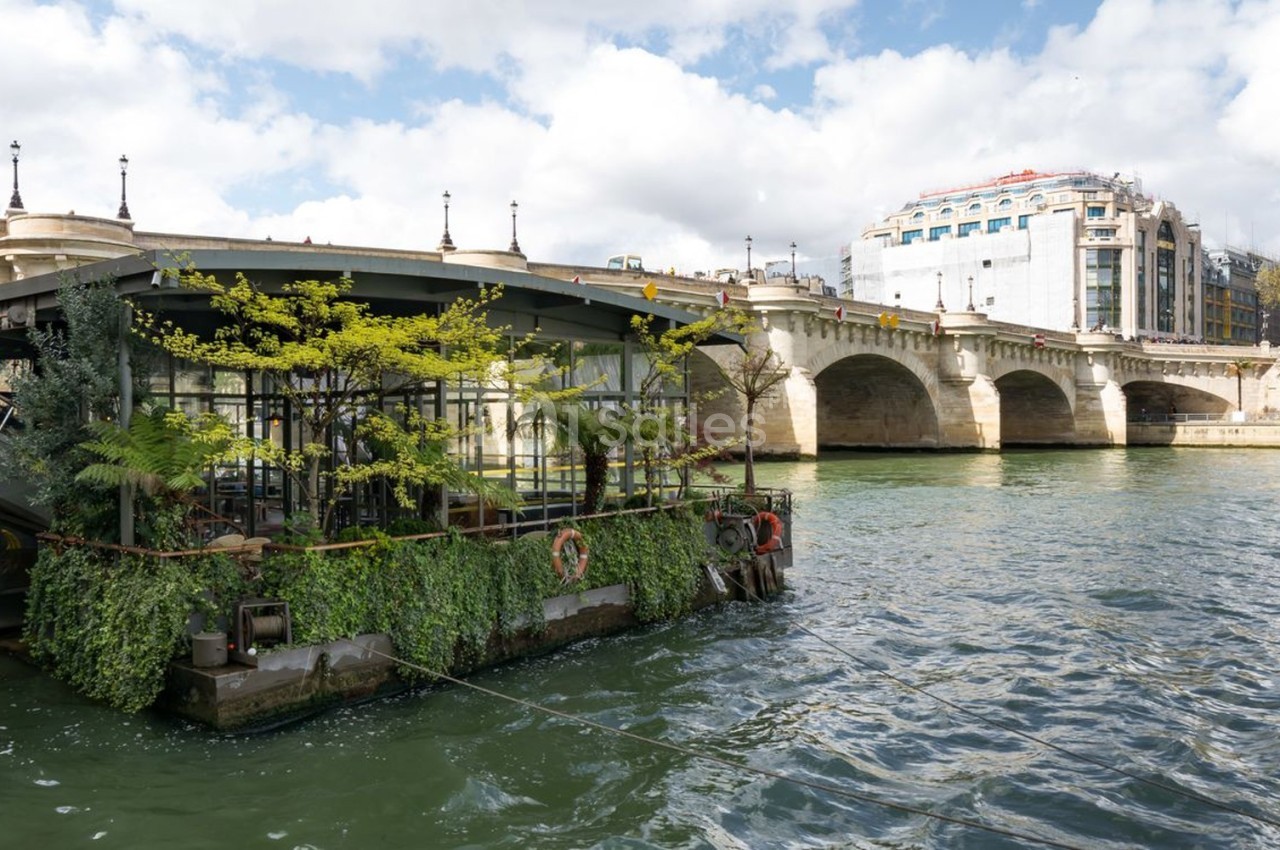Péniche végétalisée amarrée sur la Seine, près d'un pont en pierre et de bâtiments urbains en arrière-plan.