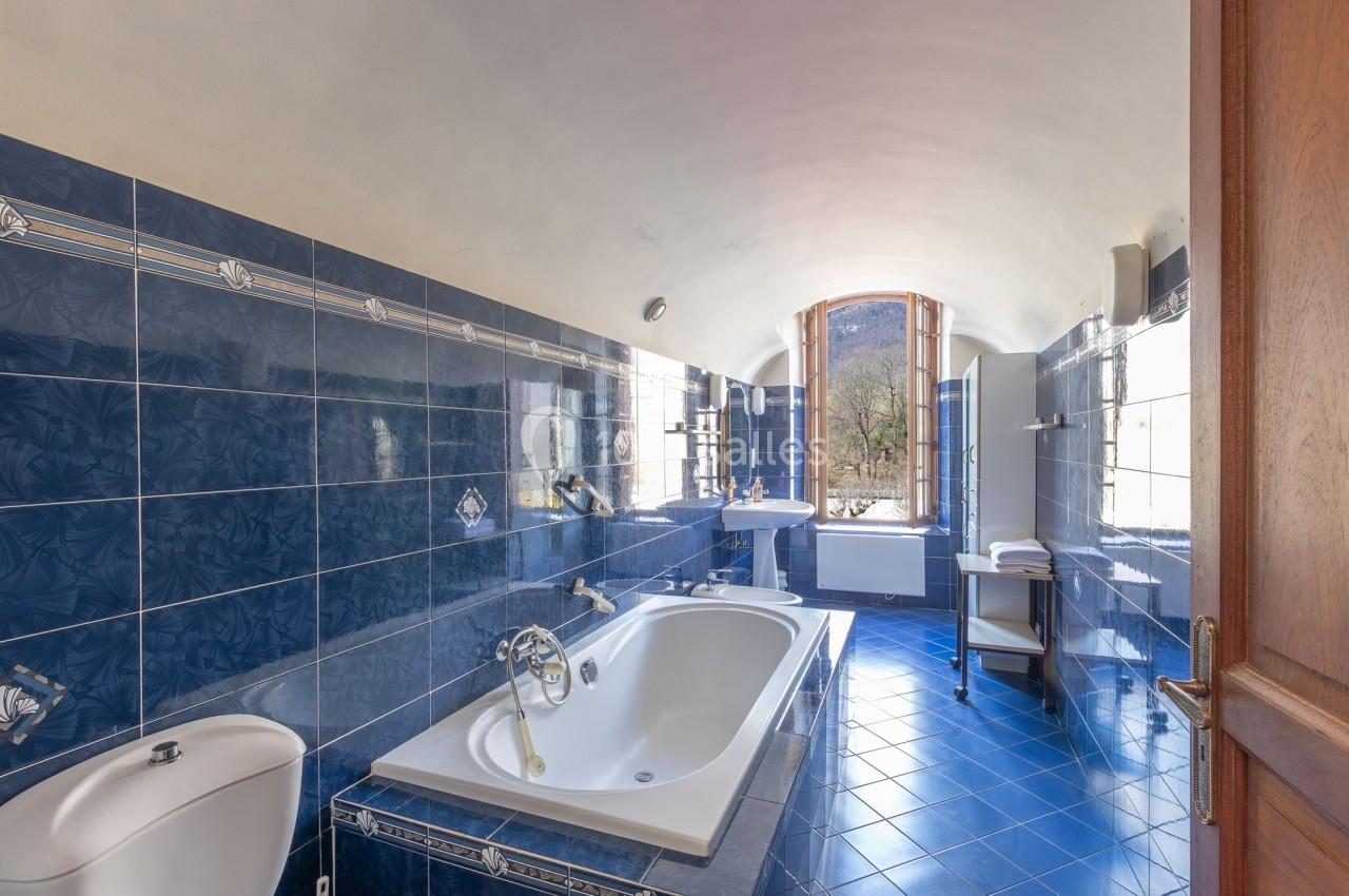 Salle de bain avec carrelage bleu, baignoire, lavabo, fenêtre ouverte sur un paysage extérieur.