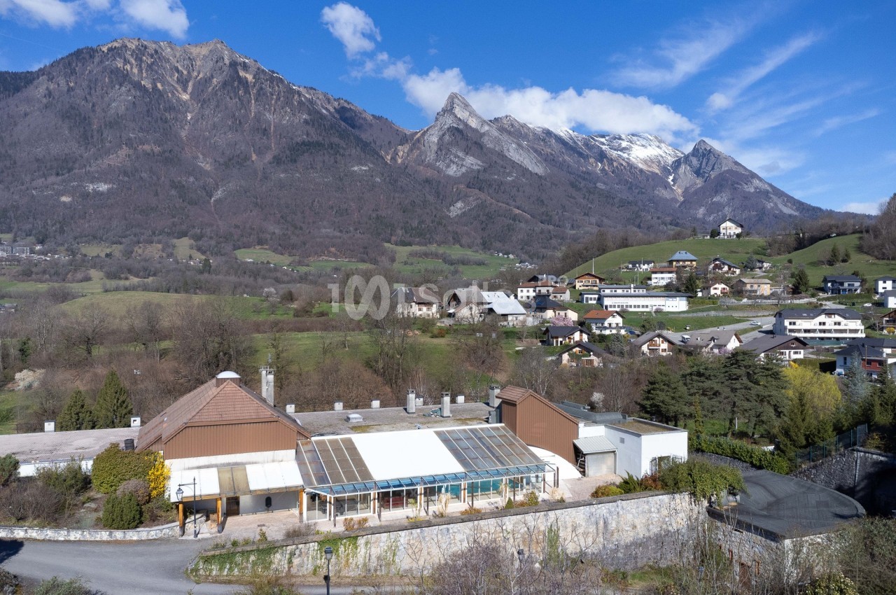 Vue d'un village alpin avec des maisons, une serre et des montagnes en arrière-plan sous un ciel bleu.