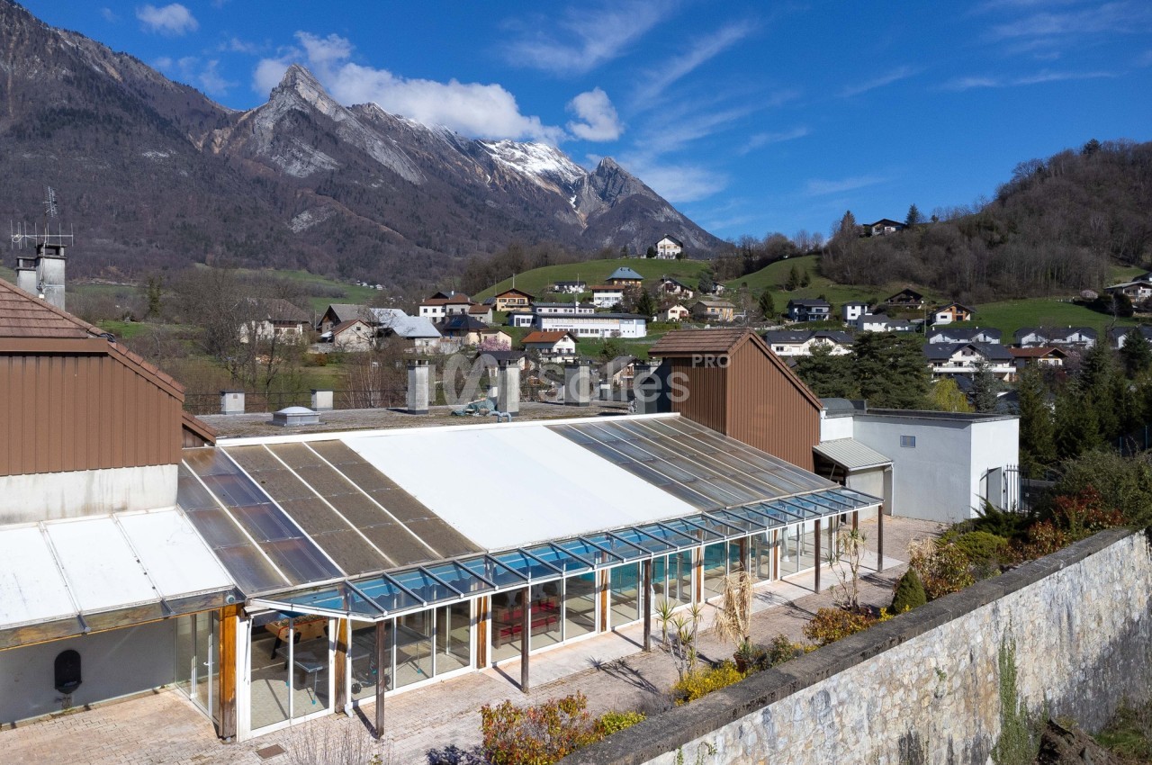 Vue d'une serre vitrée entourée de bâtiments, avec un village et des montagnes en arrière-plan sous un ciel bleu.