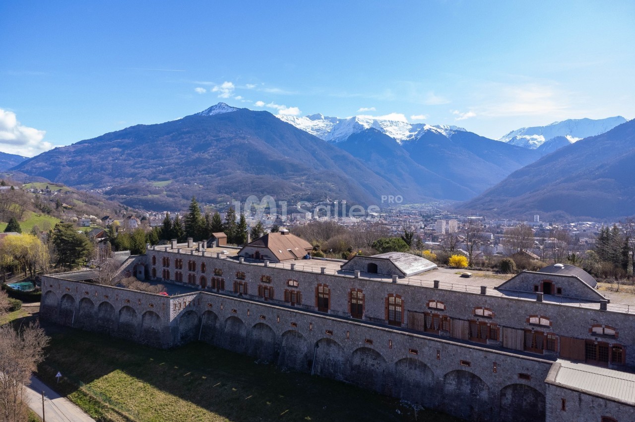 Vue aérienne d'un bâtiment historique en pierre entouré de montagnes enneigées et d'une vallée en arrière-plan.
