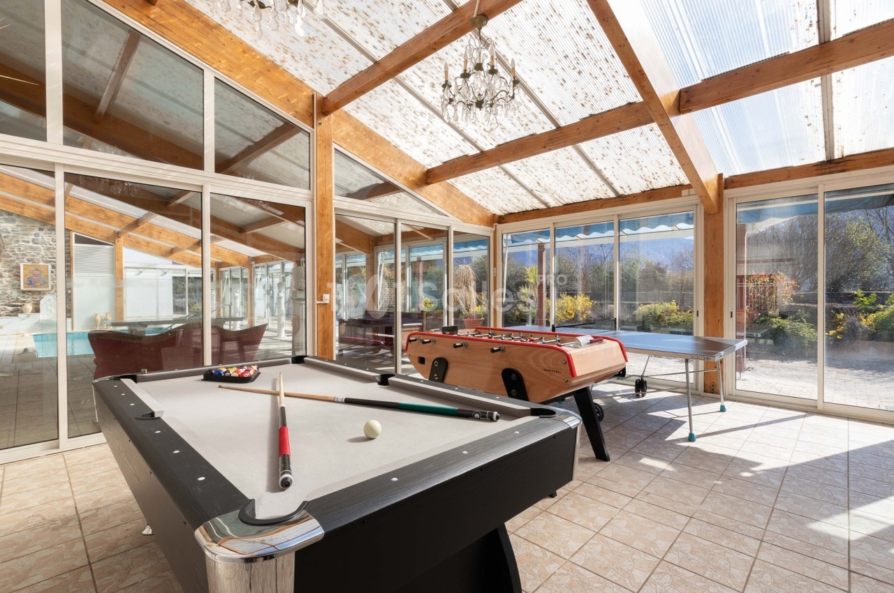 Salle lumineuse avec billard, baby-foot et table de ping-pong, entourée de baies vitrées donnant sur un jardin.