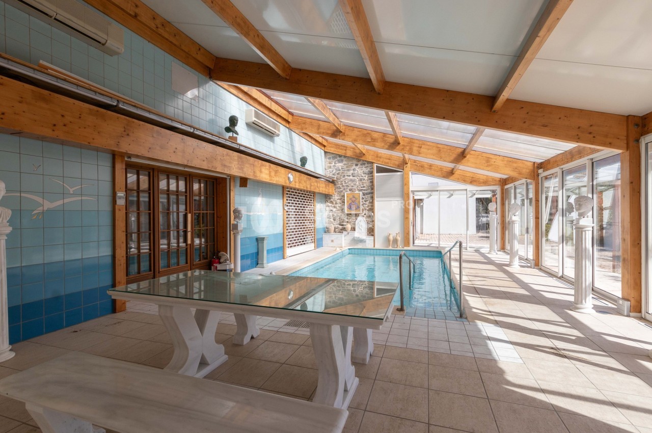 Piscine intérieure lumineuse avec toit en verre, murs en carrelage bleu, table en verre et bancs blancs.