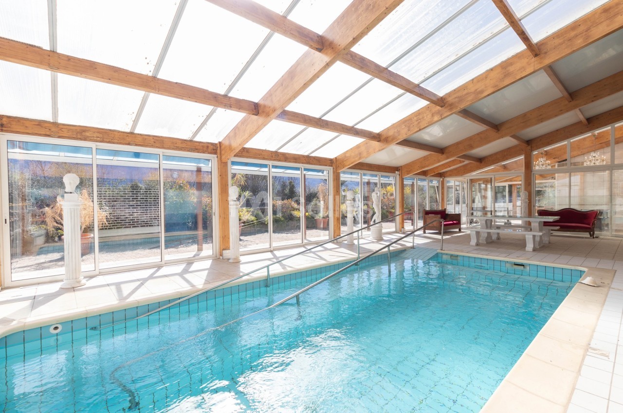 Piscine intérieure avec toit en verre, entourée de baies vitrées donnant sur un jardin ensoleillé.