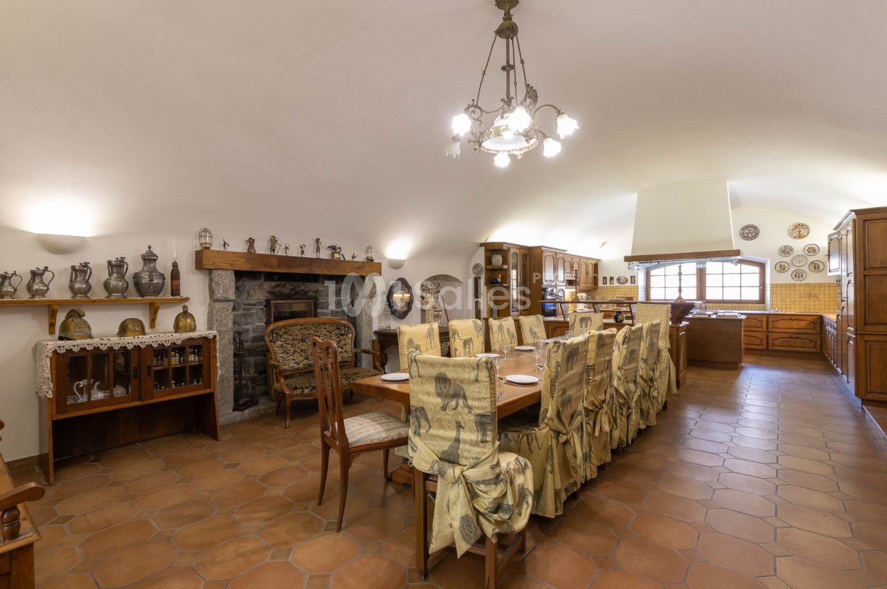 Salle à manger rustique avec table longue, chaises décorées, cheminée en pierre et cuisine ouverte en arrière-plan.