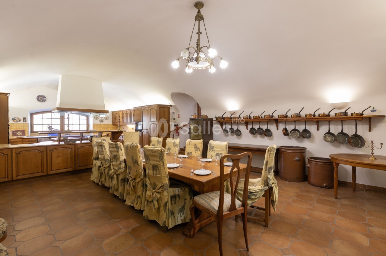 Grande cuisine rustique avec table à manger, chaises décorées, étagère murale avec poêles et carrelage au sol.