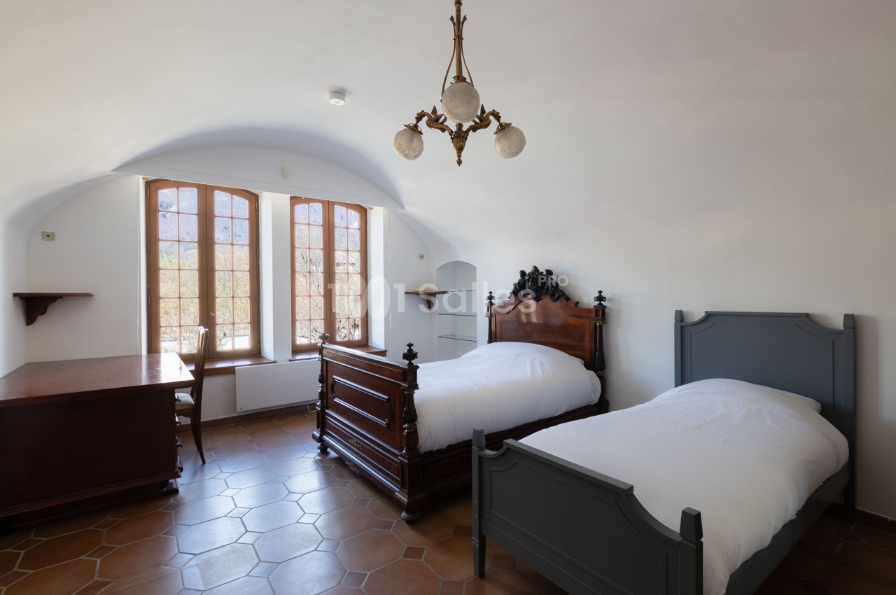Chambre lumineuse avec deux lits simples, un bureau en bois, un lustre ancien et des fenêtres donnant sur l'extérieur.