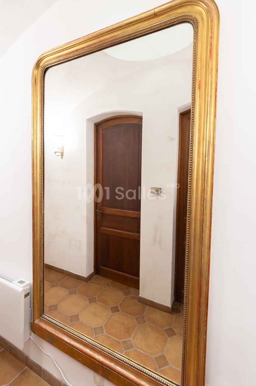 Grand miroir doré reflétant une porte en bois, un mur blanc et un sol en carreaux beige.