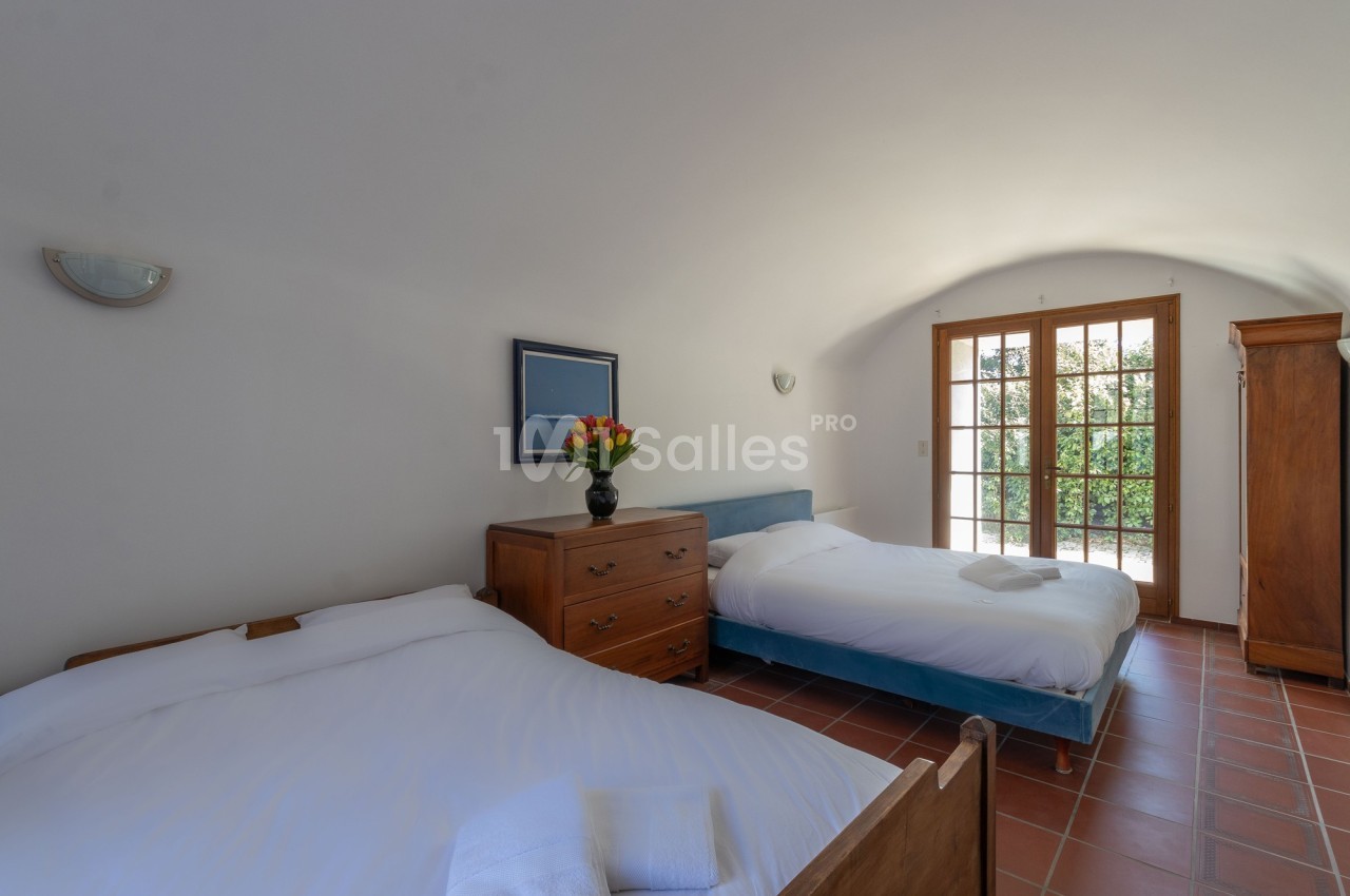 Chambre lumineuse avec deux lits, une commode, une armoire en bois et une porte-fenêtre donnant sur un jardin.
