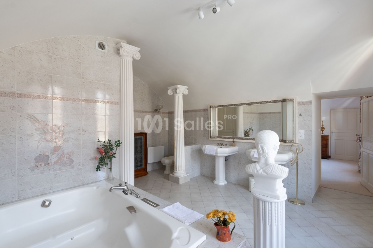 Salle de bain lumineuse avec baignoire, colonnes décoratives, miroir mural et carrelage clair.