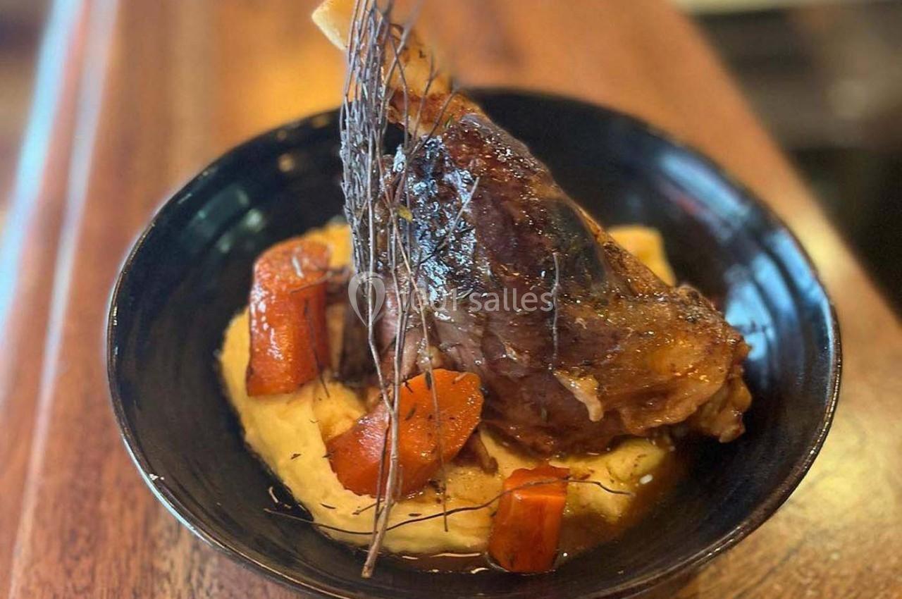 Jarret d'agneau braisé servi sur une purée crémeuse avec des morceaux de carottes, présenté dans une assiette noire.