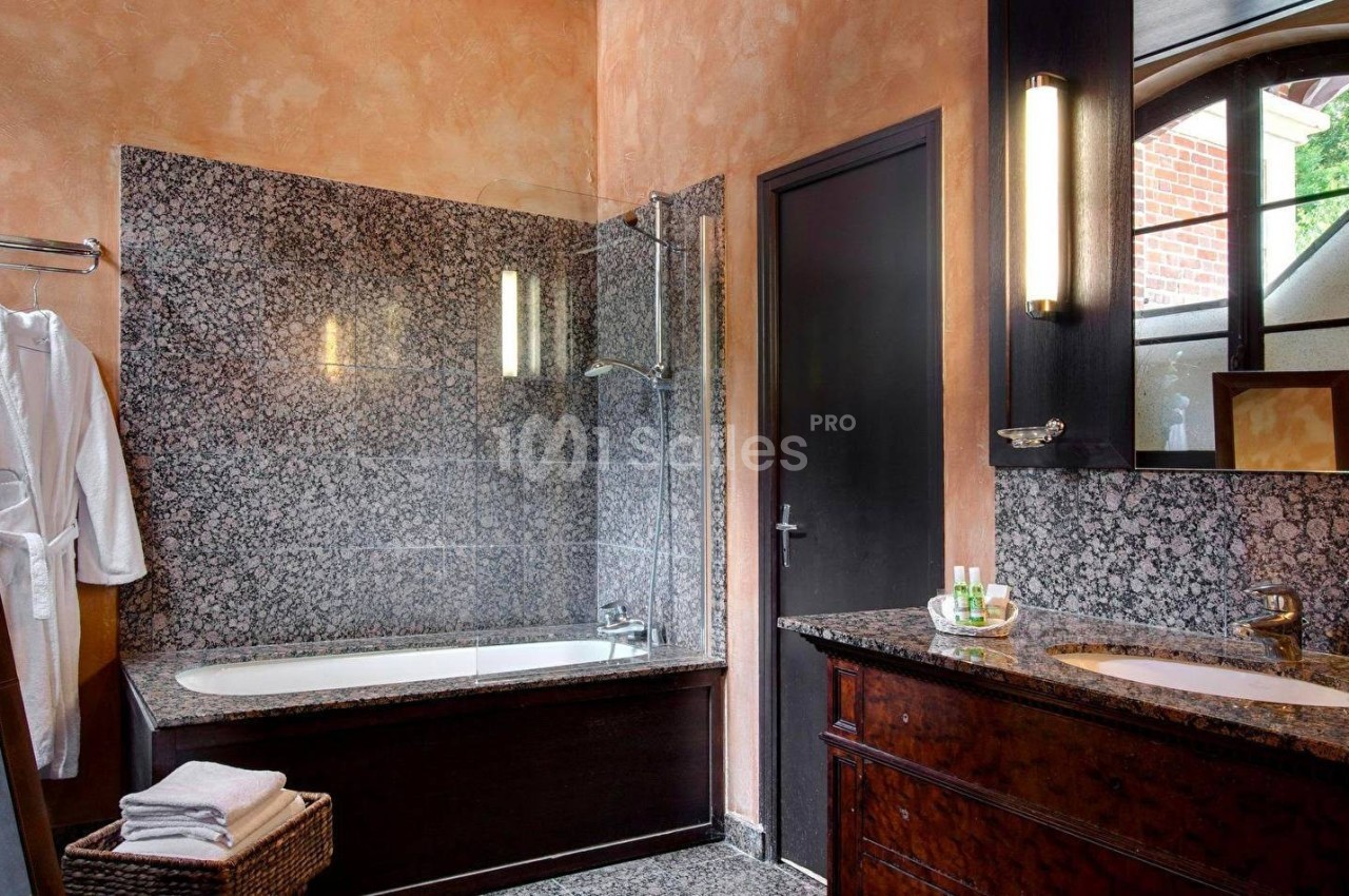 Salle de bain avec baignoire en marbre, lavabo, miroir, peignoir suspendu et fenêtre laissant entrer la lumière.