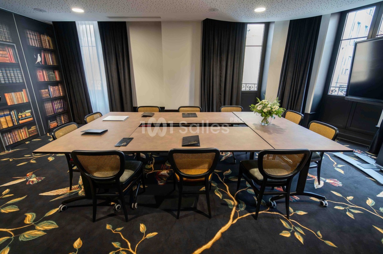 Salle de réunion lumineuse avec grande table rectangulaire, chaises en rotin, étagères de livres et décoration florale.