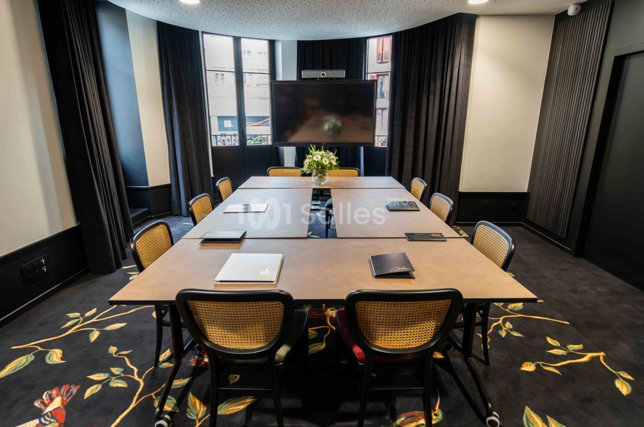 Salle de réunion avec table rectangulaire, chaises en bois, écran mural et décoration florale au sol.