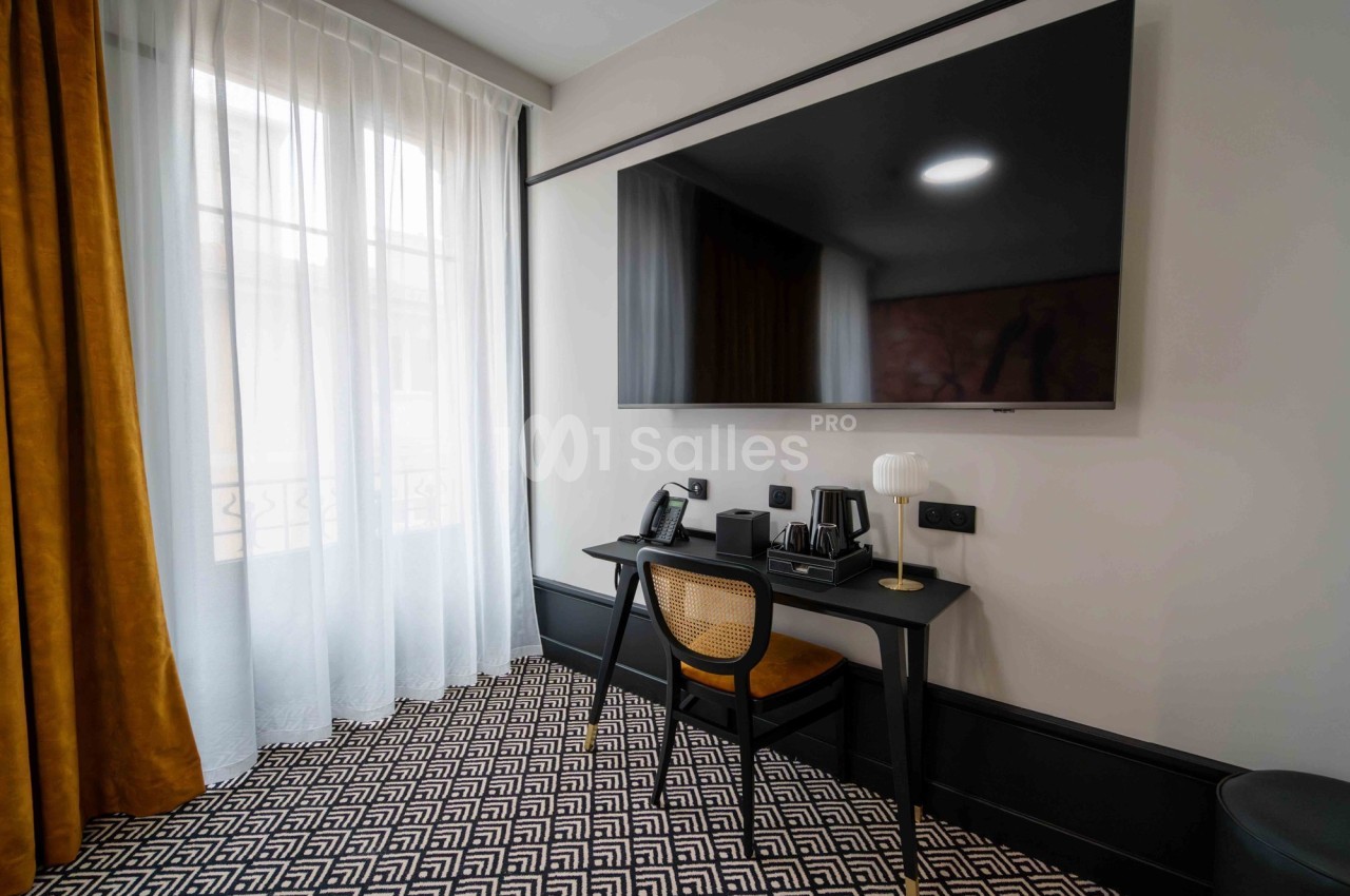 Bureau avec chaise, lampe, téléphone et plateau de courtoisie, face à un grand téléviseur mural dans une pièce lumineuse.