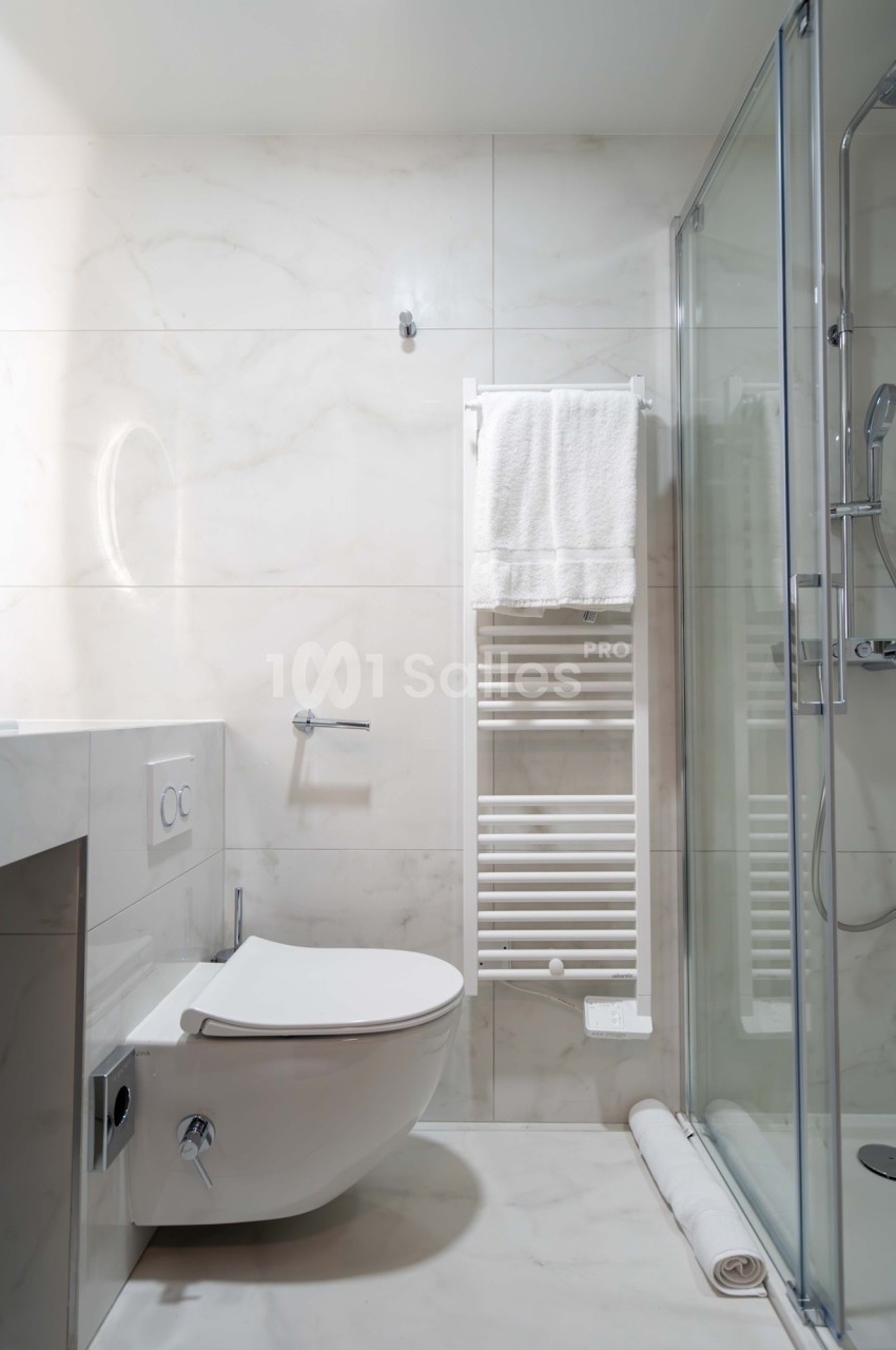 Salle de bain moderne avec toilettes suspendues, sèche-serviettes mural, douche vitrée et murs en carrelage clair.