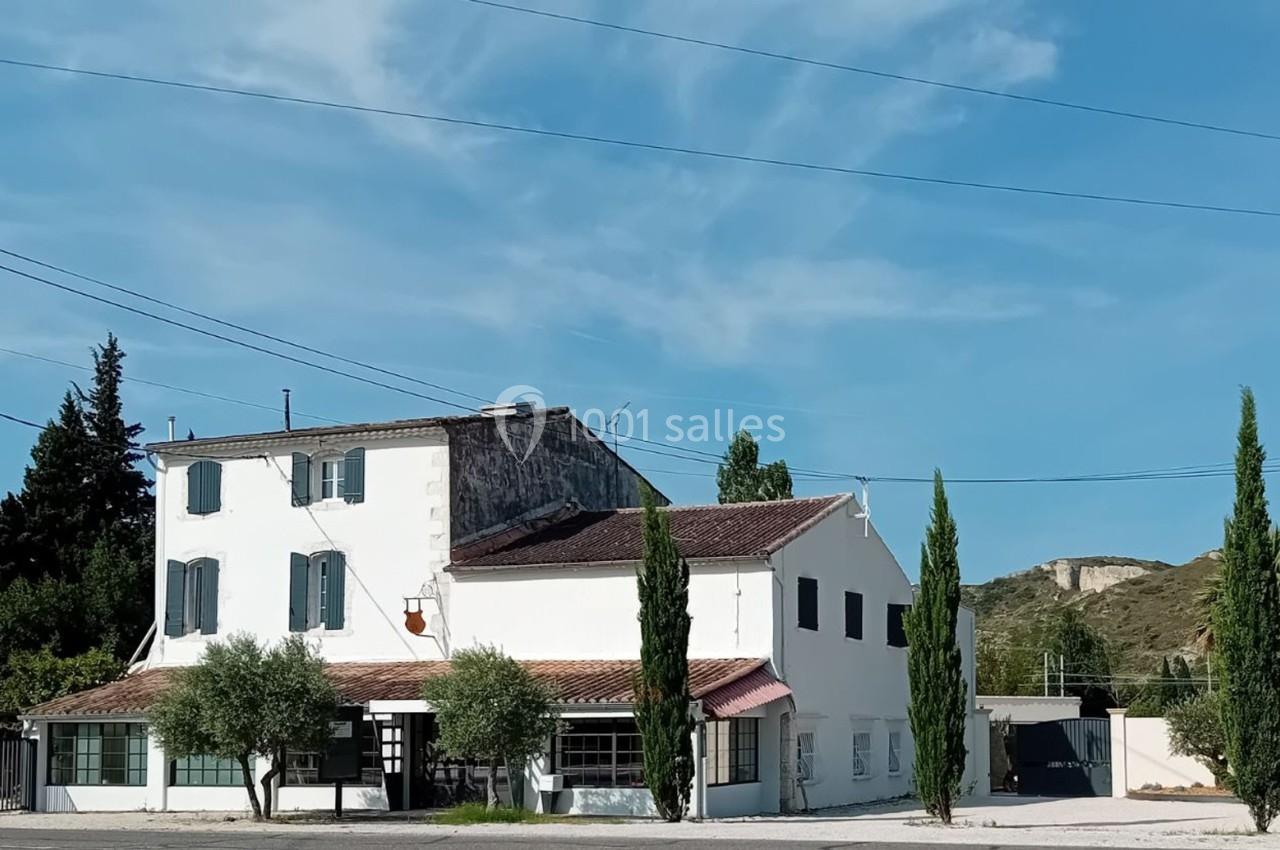 Bâtiment blanc à deux étages avec volets bleus, entouré de cyprès et d'oliviers, sous un ciel dégagé.