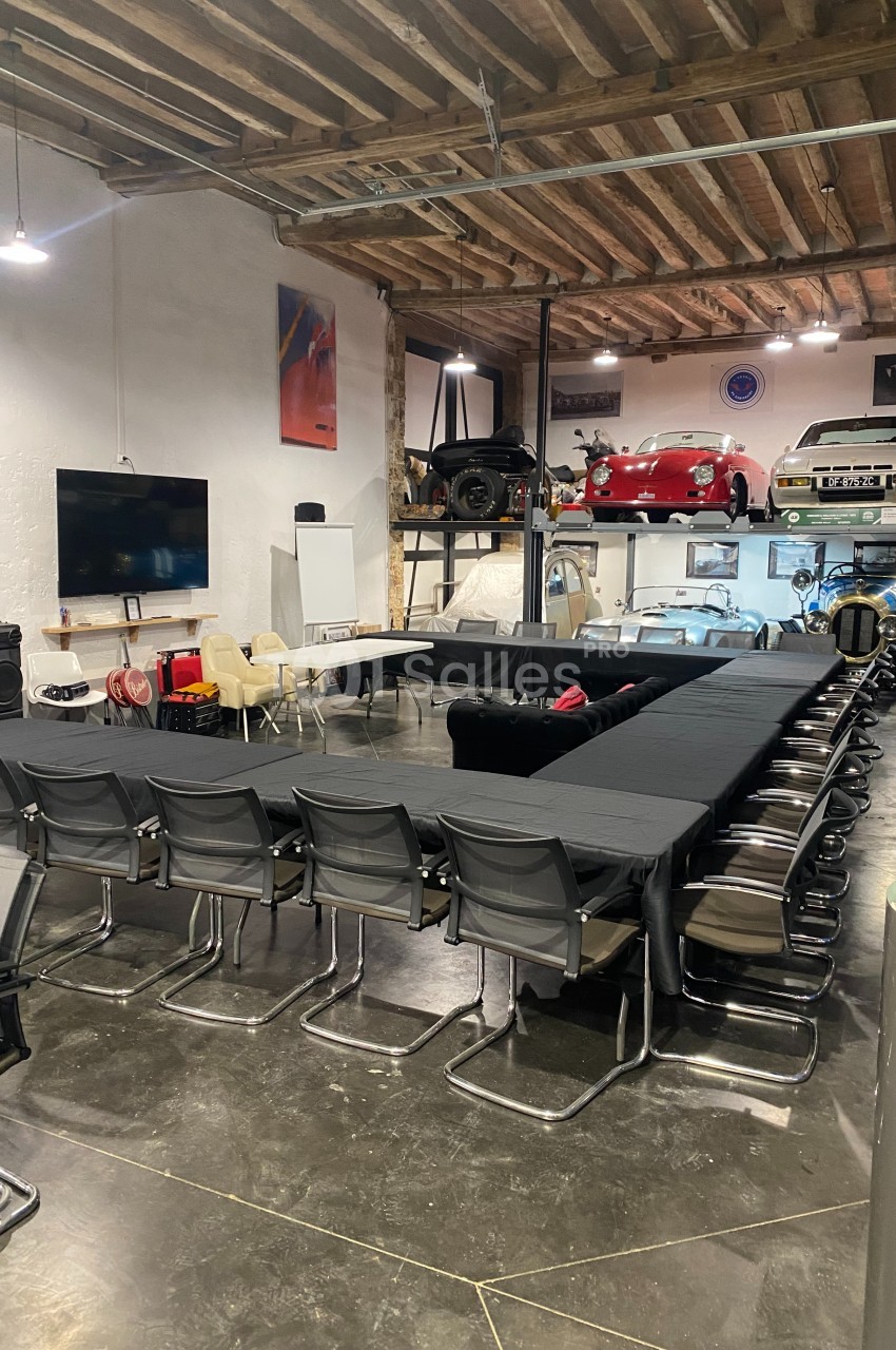 Salle de réunion aménagée dans un garage avec des voitures exposées en arrière-plan et des tables disposées en U.