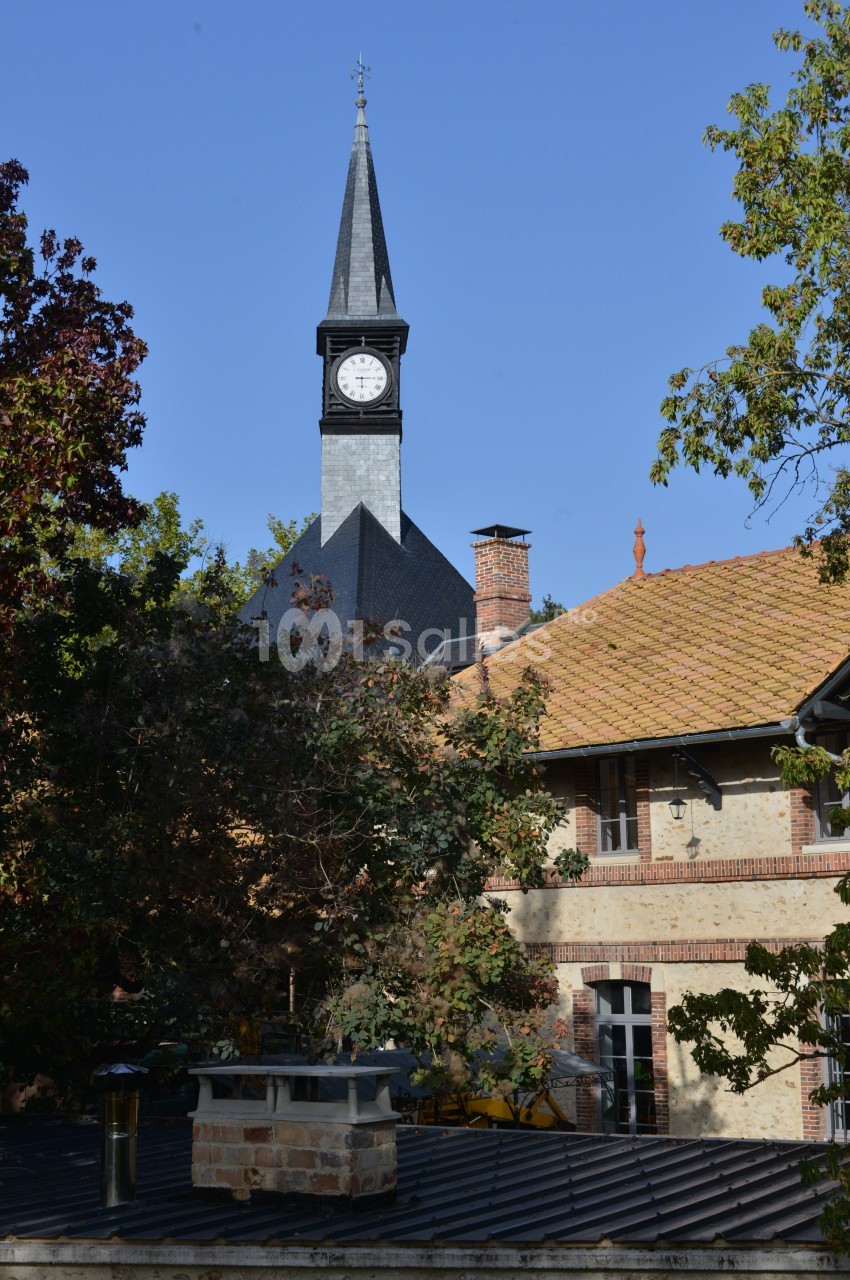 Clocher d'église avec horloge visible, entouré de bâtiments en briques et de végétation sous un ciel bleu.