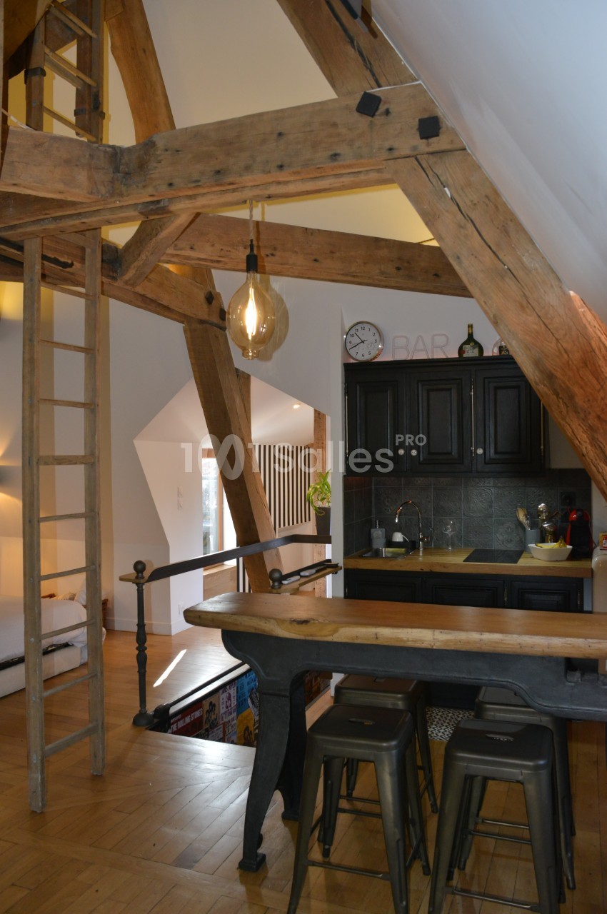 Cuisine ouverte dans un intérieur avec poutres apparentes, tabourets hauts, et éclairage suspendu.