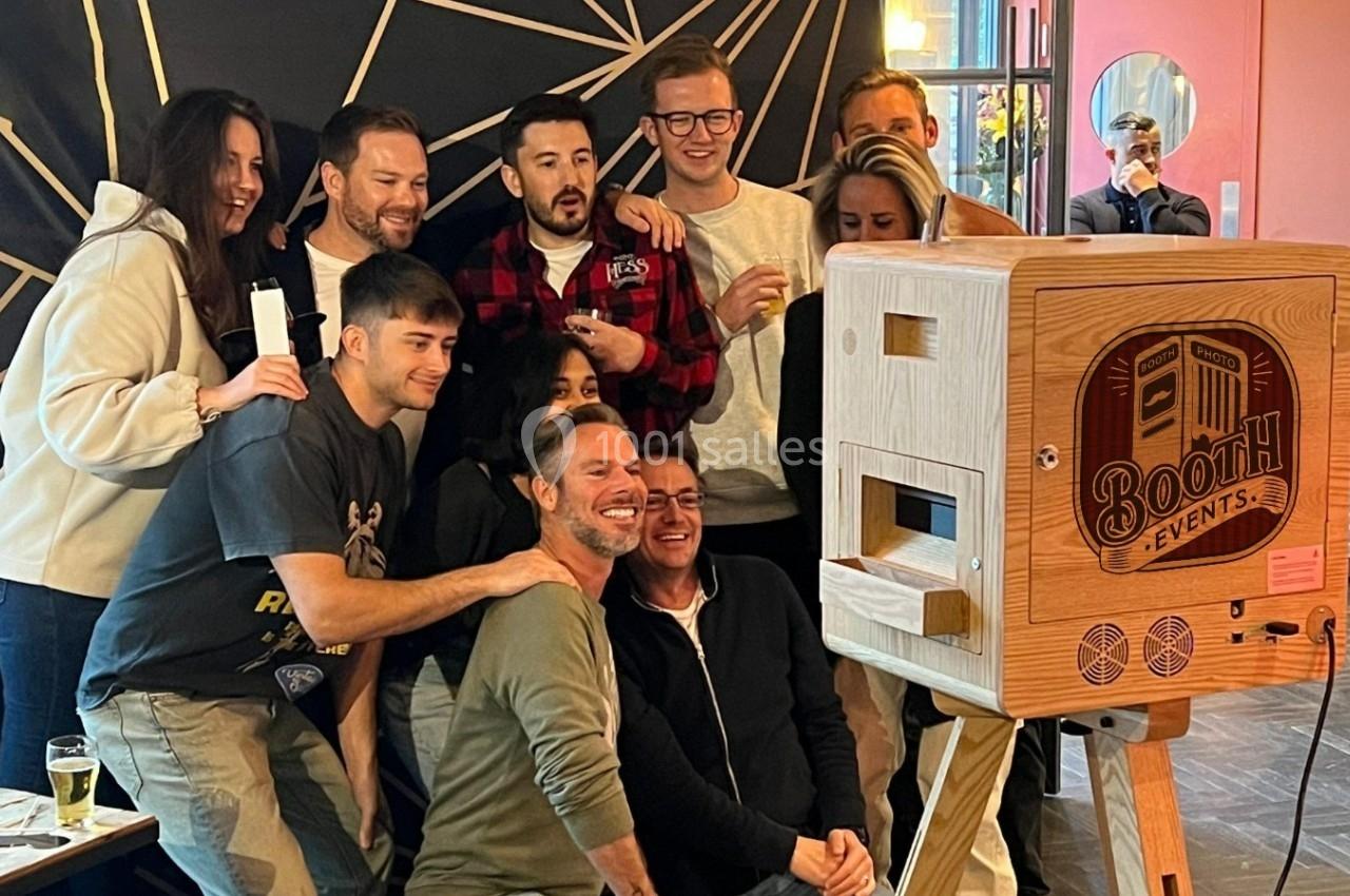 Un groupe de personnes souriantes posant devant une borne photo en bois dans un espace intérieur.