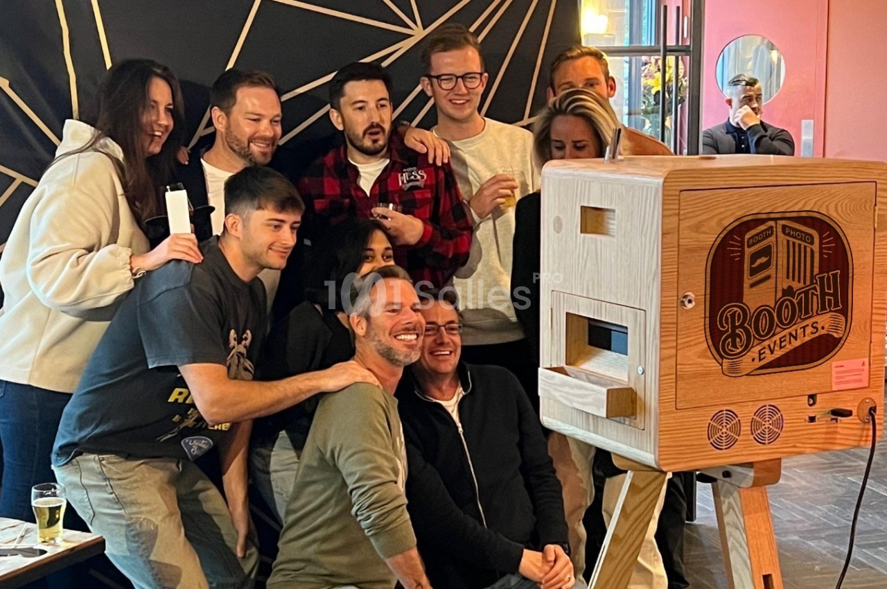 Un groupe de personnes souriantes posant devant une borne photo en bois dans un espace intérieur.
