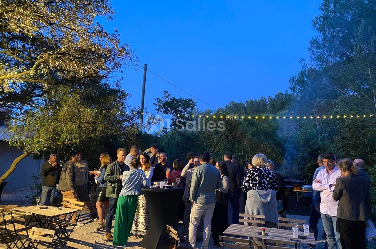 Groupe de personnes discutant dans un jardin en soirée, entouré de tables et éclairé par des guirlandes lumineuses.