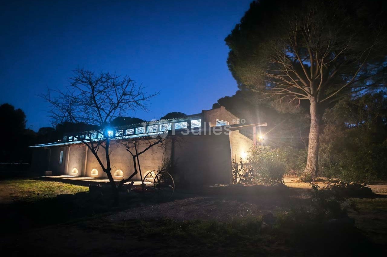 Maison moderne éclairée la nuit, entourée d'arbres et d'un jardin, avec un ciel sombre en arrière-plan.