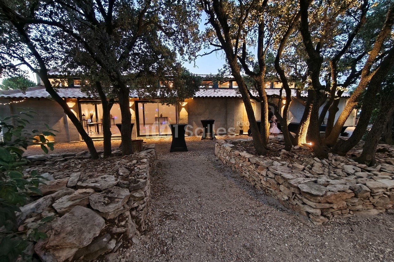 Chemin en gravier bordé de murets en pierre menant à un bâtiment éclairé entouré d'arbres au crépuscule.