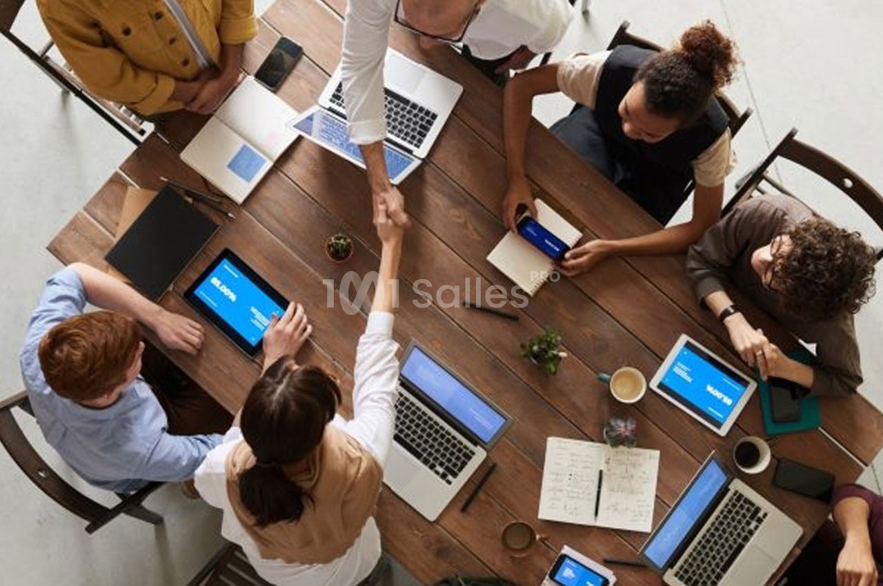 Personnes collaborant autour d'une table avec ordinateurs, tablettes, carnets et une poignée de main au centre.