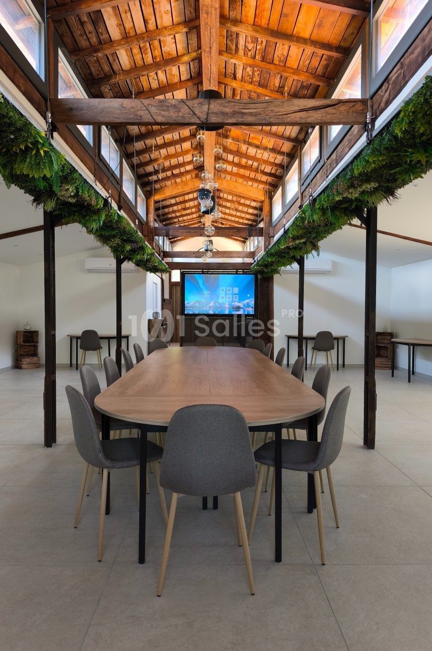 Salle de réunion lumineuse avec une grande table en bois entourée de chaises grises, sous un toit en bois avec plantes…
