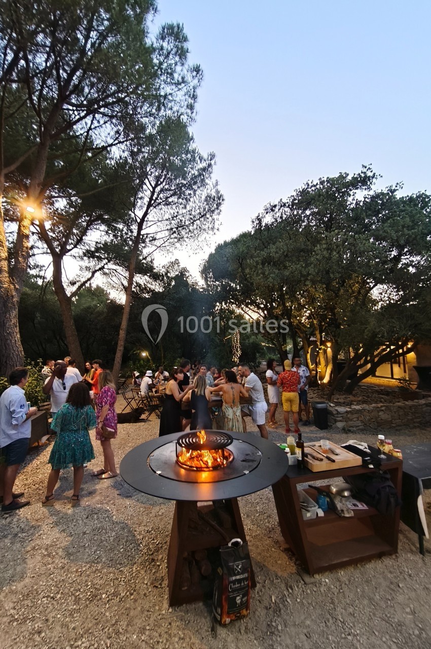 Groupe de personnes réunies en extérieur autour d'un brasero allumé, entourées d'arbres et de tables.