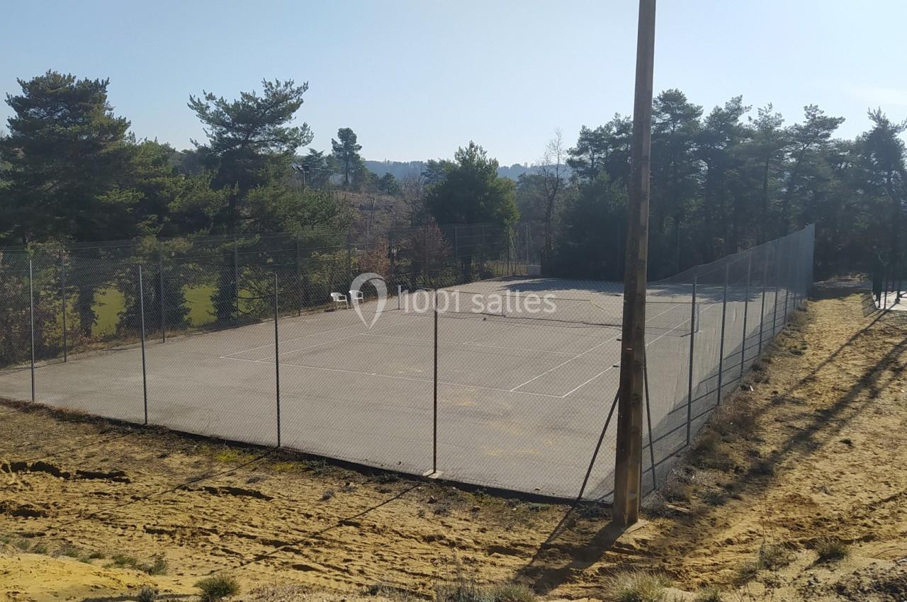 Terrain de tennis extérieur entouré de grillages, situé dans un environnement boisé sous un ciel dégagé.
