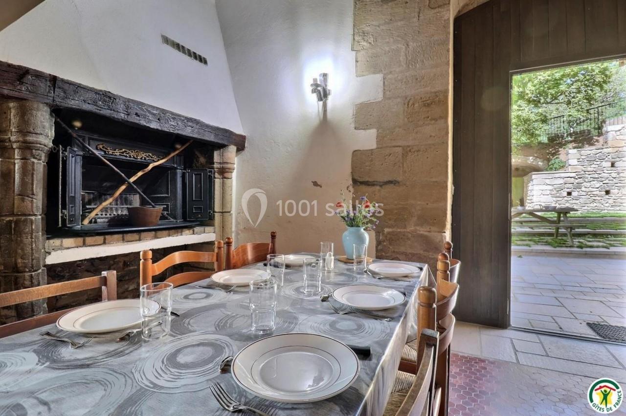 Table dressée avec vaisselle et verres dans une salle à manger rustique, cheminée ancienne et vue sur une cour extérieure.