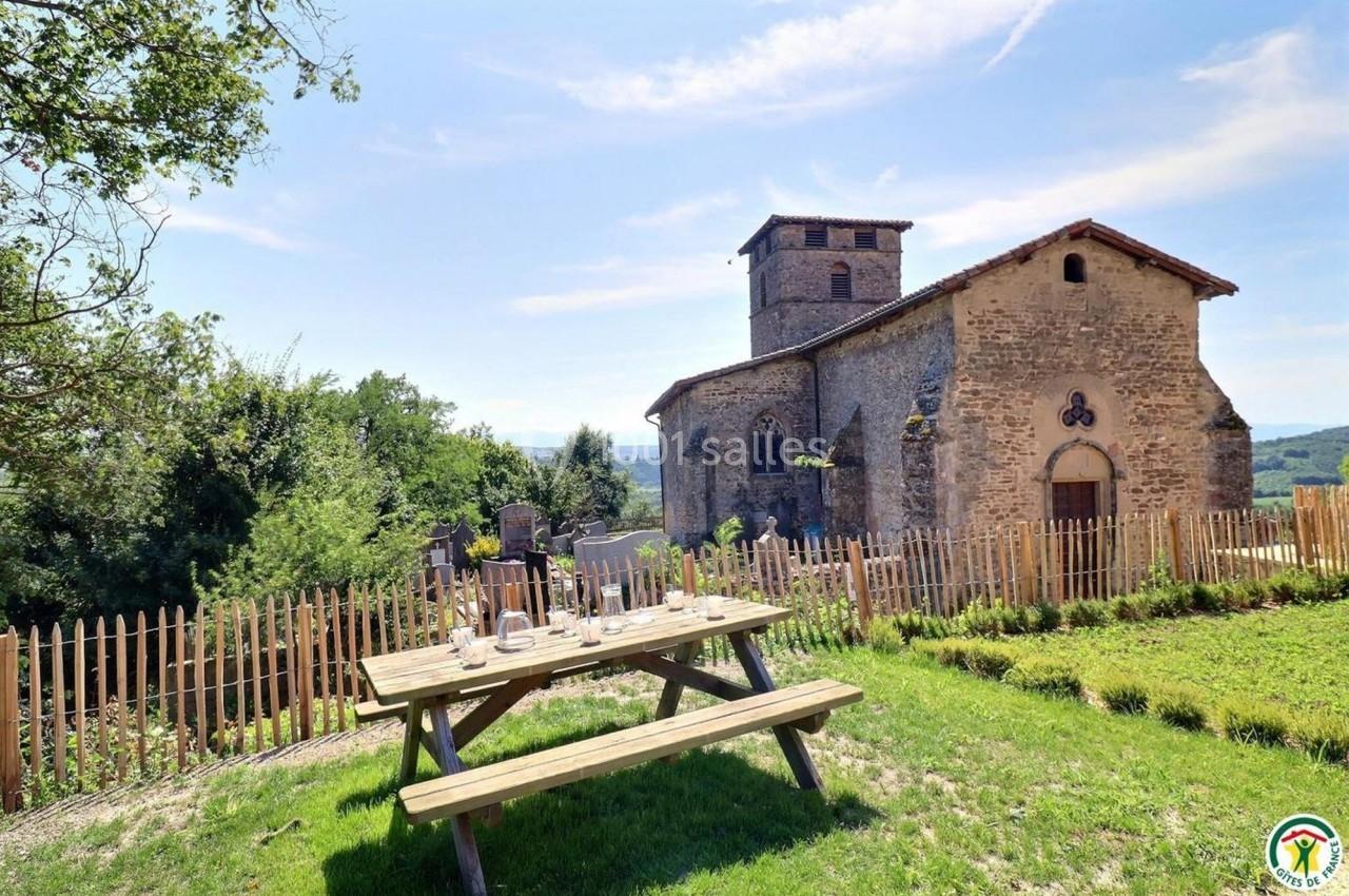 Table de pique-nique en bois devant une petite église en pierre entourée de verdure et d'une clôture en bois.