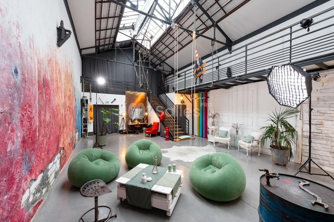 Loft lumineux avec verrière, mobilier éclectique, poufs verts, escalier métallique et décorations artistiques.