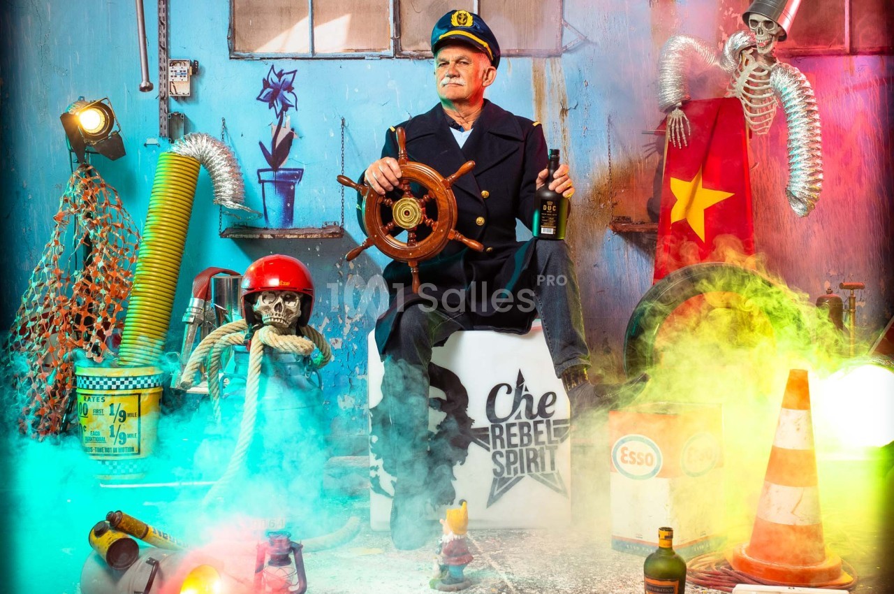 Un homme déguisé en marin tient un gouvernail dans un décor coloré et chaotique avec divers objets insolites.