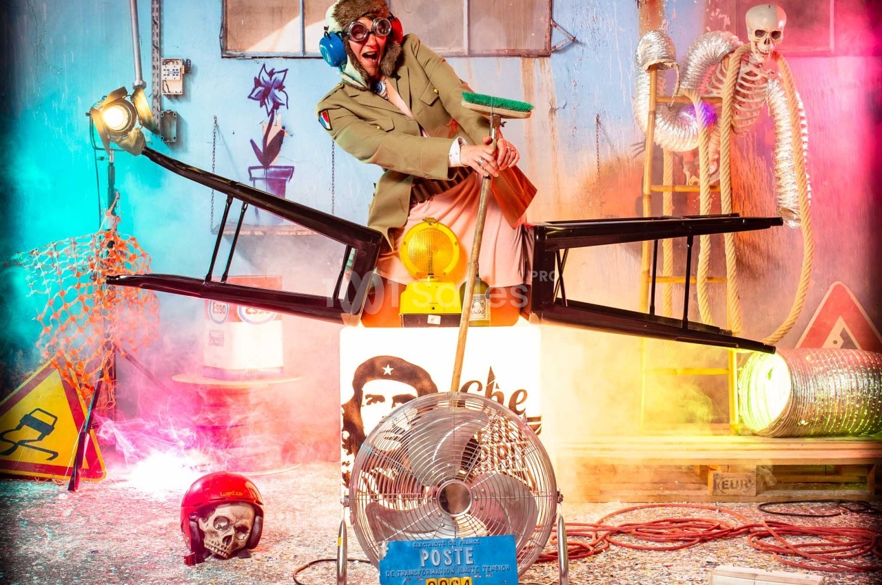 Un homme déguisé en pilote sur une chaise montée sur un ventilateur, entouré de décors colorés et d'objets insolites.