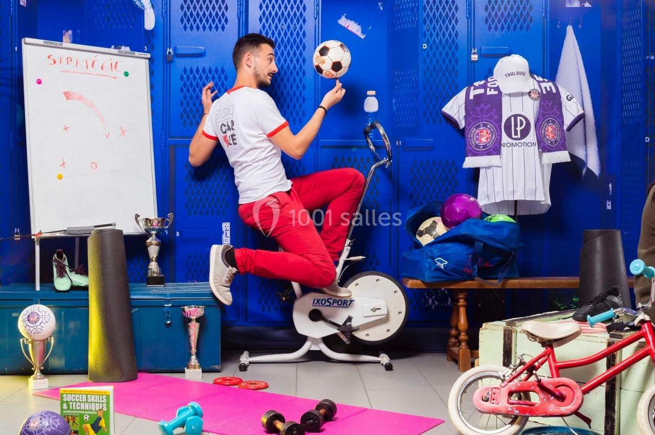 Un homme en tenue sportive jongle avec un ballon de football sur un vélo d'appartement dans un vestiaire coloré.