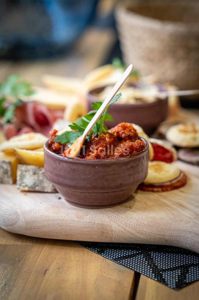 Bol de tartinade rouge avec une cuillère en bois, entouré de charcuteries, fromages et pains sur une planche en bois.