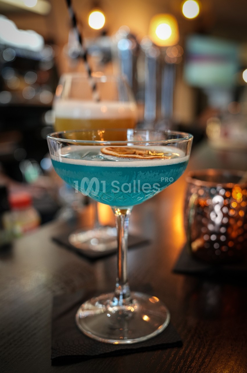 Cocktail bleu garni d'une tranche d'agrume séché, posé sur un comptoir de bar avec un autre verre en arrière-plan.