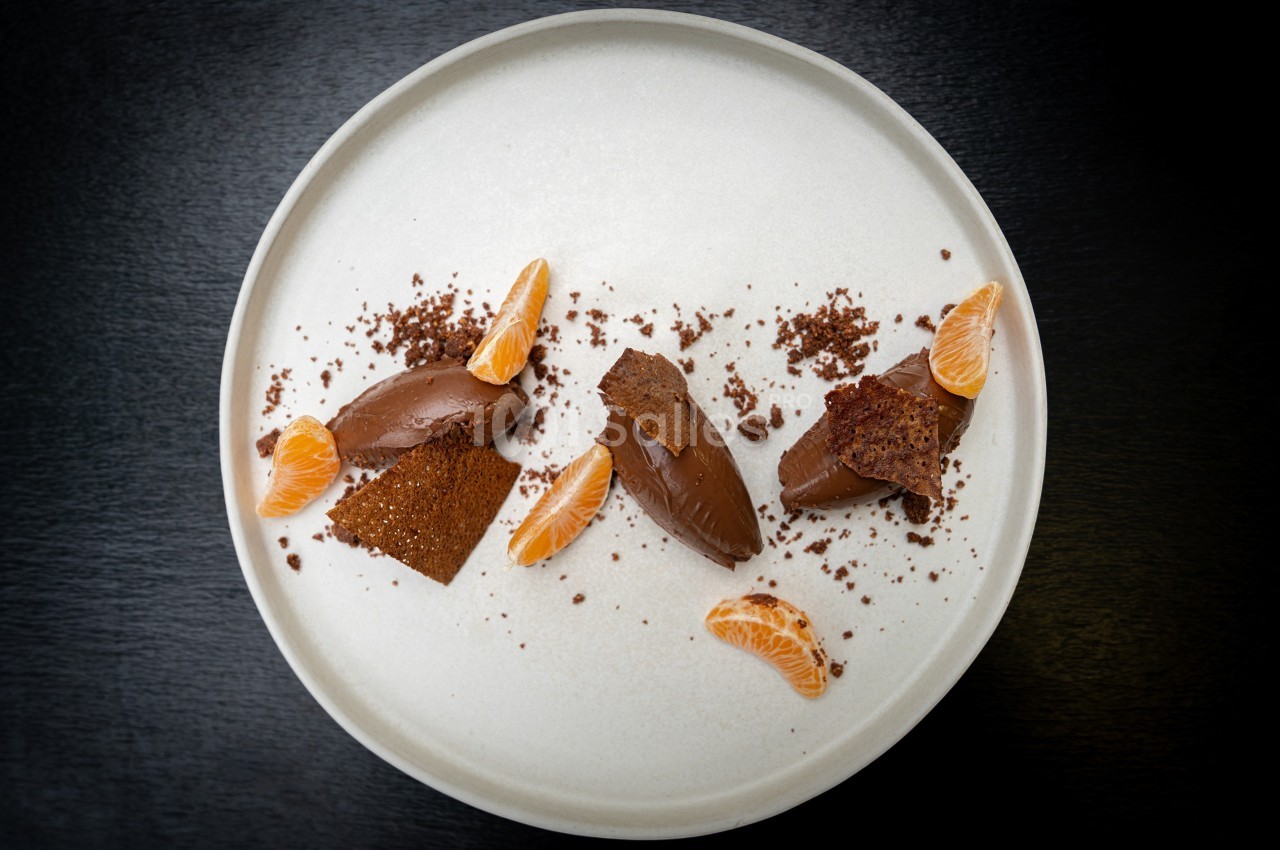 Dessert composé de quenelles de mousse au chocolat, segments de mandarine et éclats de biscuit sur une assiette blanche.