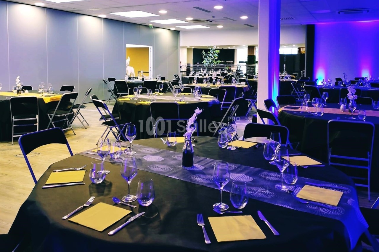 Salle de réception aménagée avec des tables rondes dressées, nappes noires, serviettes jaunes et éclairage tamisé.