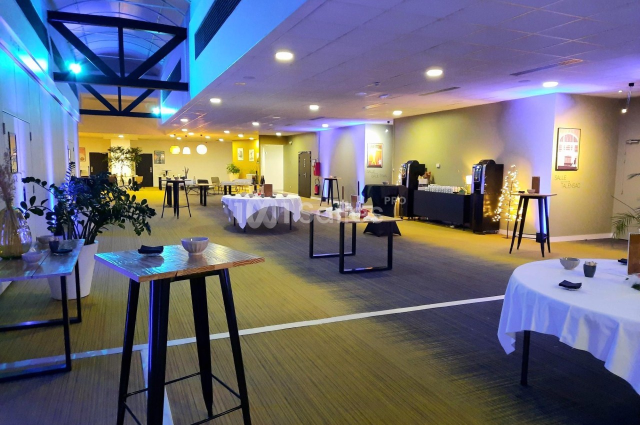 Salle lumineuse aménagée pour un événement, avec tables hautes, nappes blanches et buffet en arrière-plan.